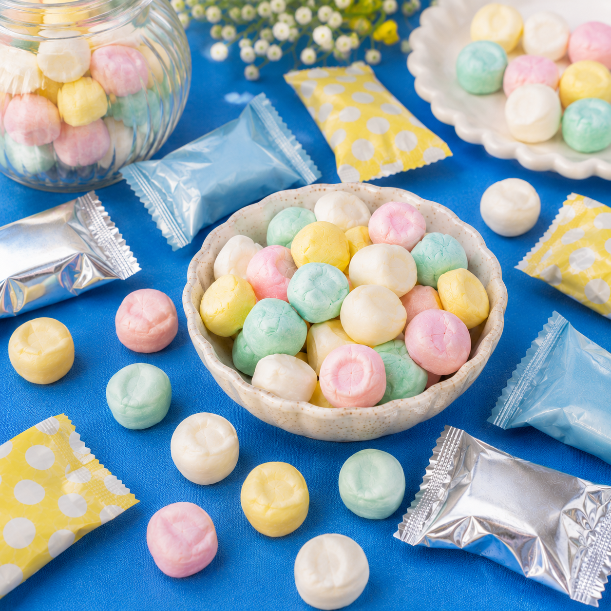 Pastel buttermint candies in pink, yellow, mint green, and white displayed in a scalloped bowl on a bright blue tabletop, with wrapped candies scattered around.