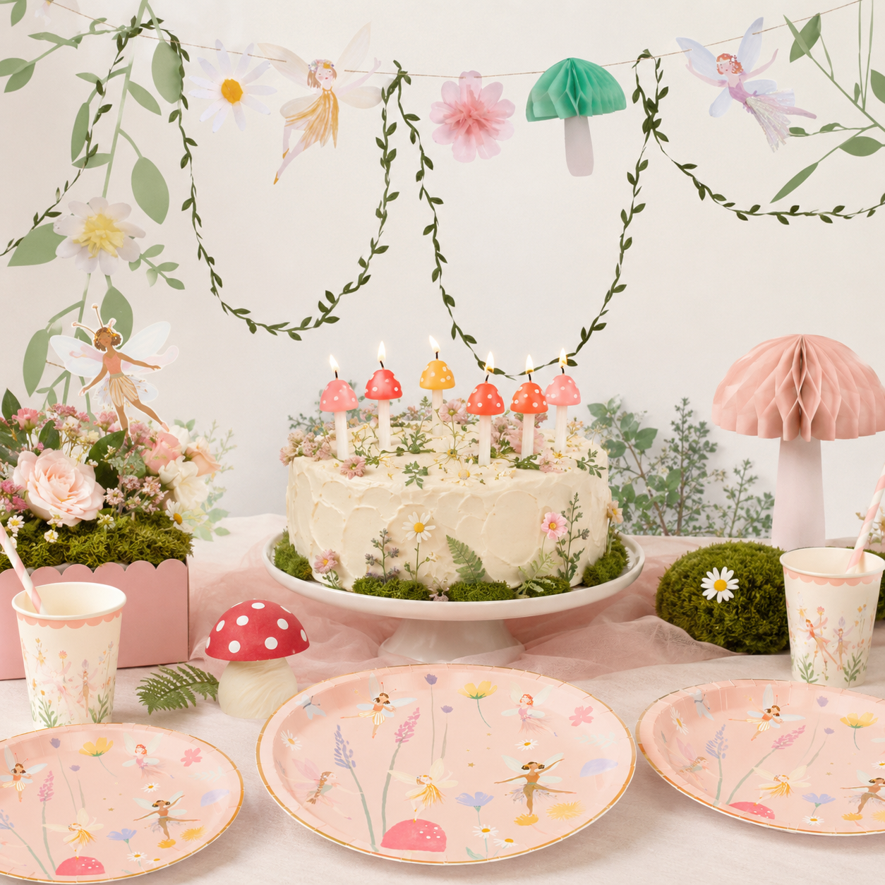 Fairy garden birthday table with pastel fairy plates and cups, a floral cake with mushroom candles, fairy garland, leafy vines, flowers, moss, and mushroom decorations.