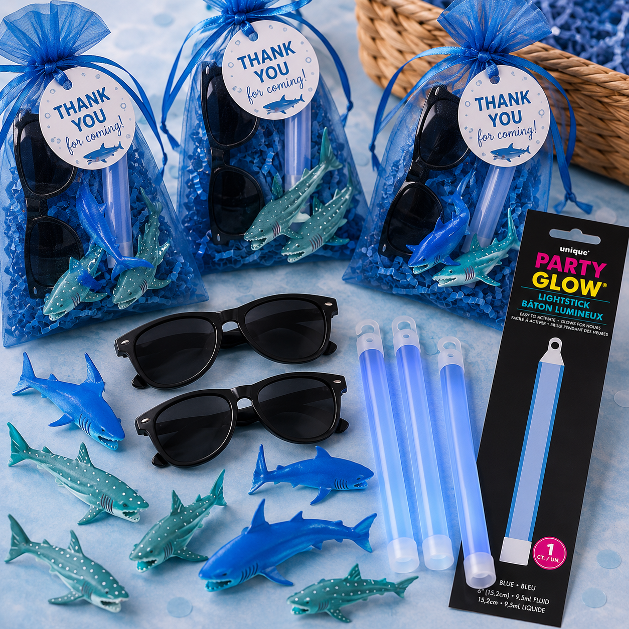 Shark party favors with blue organza bags, thank-you tags, toy sharks, black sunglasses, blue glow sticks, shredded filler, and shark-themed party favors.