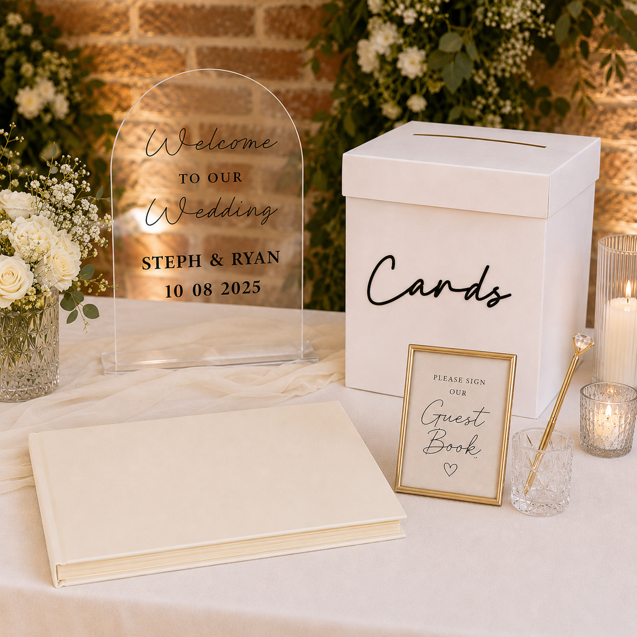 Wedding welcome table with an acrylic welcome sign, white cards box, ivory landscape guest book, white florals, candles, gold pen, and soft brick backdrop.