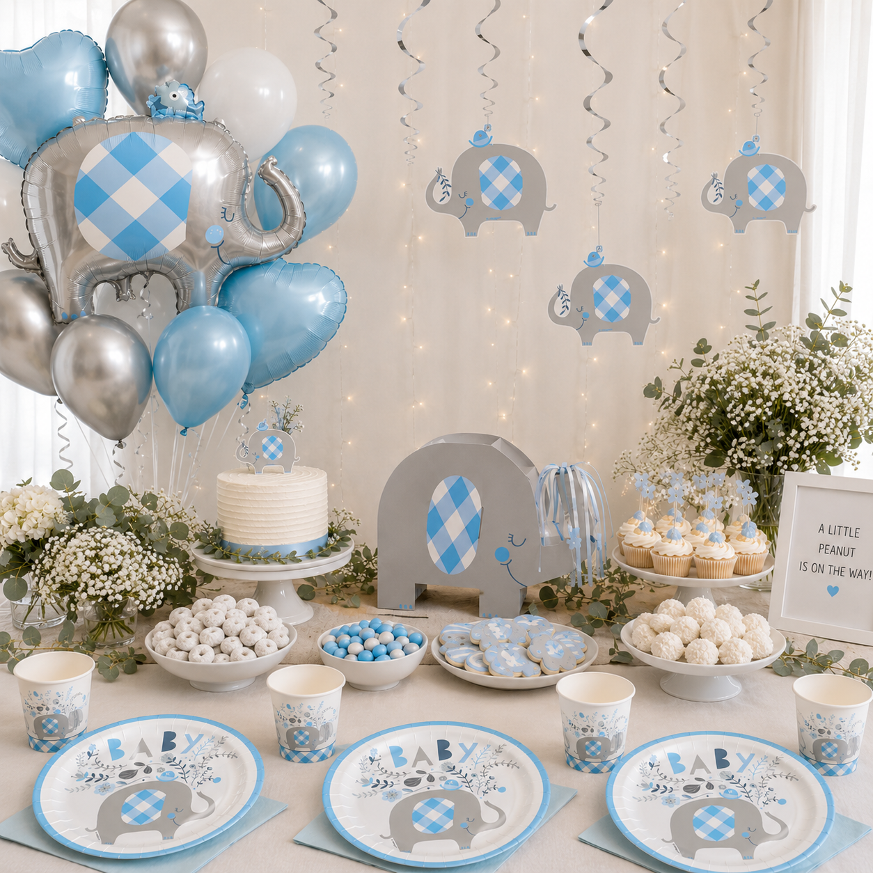 Blue elephant baby shower setup with matching plates and cups, silver and blue balloons, elephant cutouts, white floral arrangements, cake, cupcakes, cookies, candy, and “A Little Peanut Is On the Way” sign.