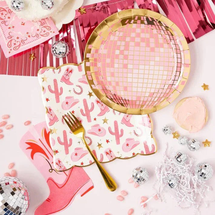 Pink western party setup with disco ball plate, cowgirl boot and cactus napkins, gold fork, pink fringe backdrop, mini disco balls, gold star confetti, and pink frosted cookie.