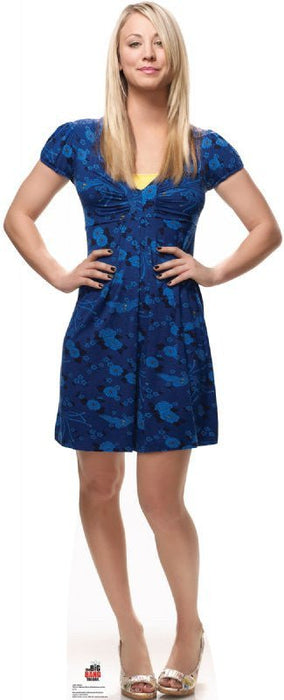 Woman with long blonde hair wearing a short-sleeved blue floral dress with gathered neckline, standing with hands on hips against a white background.