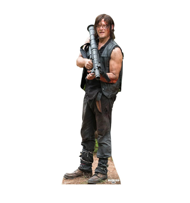 Life-size cardboard standup of a man with shoulder-length brown hair wearing a sleeveless black vest and dark pants, holding a large gray rocket launcher across his body.