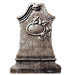 Gray stone tombstone standup with carved relief of a grinning skull holding a round bomb or sphere, topped with decorative curved elements and a textured surface.