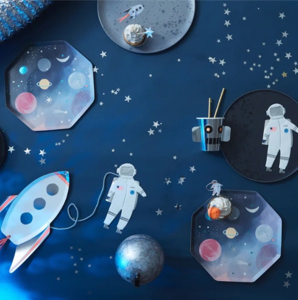 Space-themed paper products including astronaut napkins, rocket plates, and planet plates on a blue background with stars.