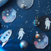 Space-themed paper products including astronaut napkins, rocket plates, and planet plates on a blue background with stars.