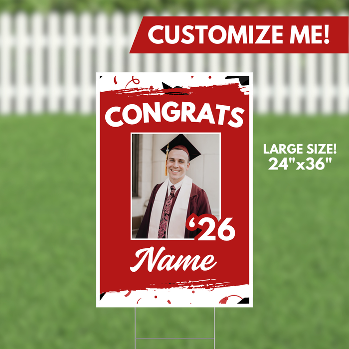 Red and white graduation yard sign, 24 by 36 inches, with "CONGRATS" text, a customizable photo frame, class year "26," and name placeholder on a white fence and grass background.