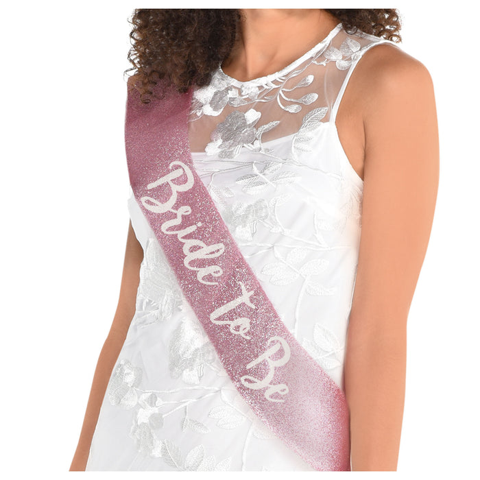 Pink glitter Bride To Be sash worn across a model’s shoulder, featuring metallic script lettering on a bridal shower accessory.