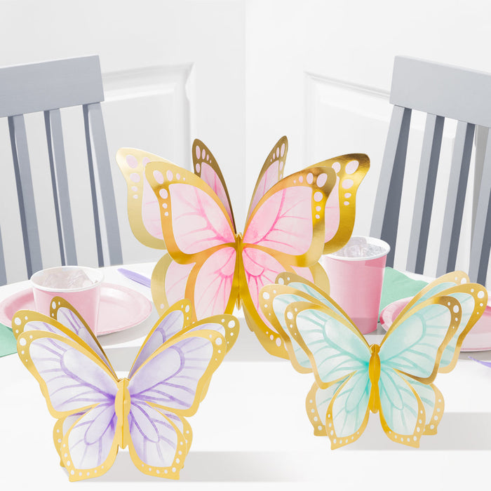 3D Butterfly Shimmer centerpieces in pastel pink, purple, and teal with gold borders displayed on a party table with pink cups and plates.
