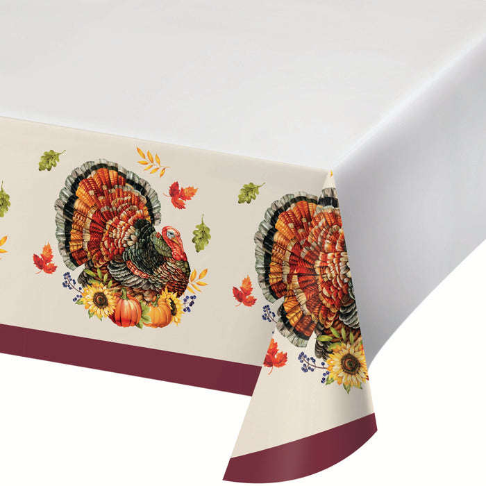 A holiday tablecloth with a cream background, featuring a large autumn turkey design, sunflowers, pumpkins, and scattered fall leaves. The bottom edge has a solid burgundy stripe.