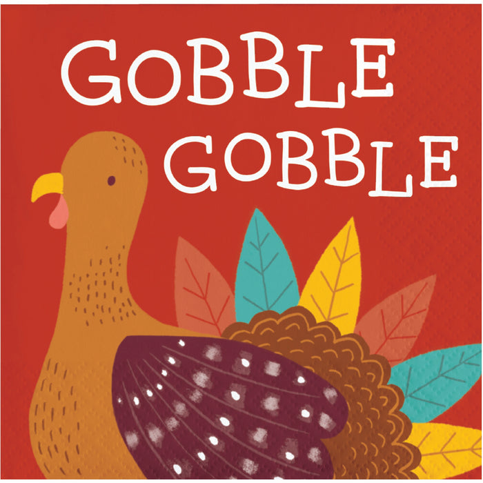 A square napkin featuring the words 'GOBBLE GOBBLE' in large white hand-drawn letters over a red background, with a stylized brown turkey and colorful autumn leaves below.