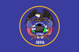 Blue flag featuring Utah's state seal with a golden eagle, beehive, and "1896" text, mounted on a wooden stick.