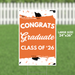 Large orange and white yard sign reading "CONGRATS Graduate CLASS OF '26" with black graduation caps and orange confetti decorations, measuring 24 inches by 36 inches.