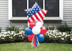 Patriotic yard sign featuring a large star-shaped balloon with white stars on blue and red and white stripes, topped with red, white, and blue balloons arranged below and ribbon streamers.