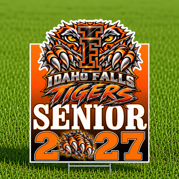 A rectangular yard sign with a Idaho Falls Tigers senior 2027 graphic, displayed on a metal stake in a green grass field. The sign features a tiger head logo, the text 'IDAHO FALLS TIGERS', 'SENIOR', and '2027' in bold orange, white, and black lettering.