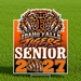 A rectangular yard sign with a Idaho Falls Tigers senior 2027 graphic, displayed on a metal stake in a green grass field. The sign features a tiger head logo, the text 'IDAHO FALLS TIGERS', 'SENIOR', and '2027' in bold orange, white, and black lettering.