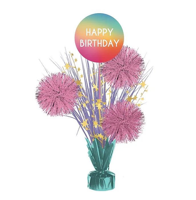 Birthday spray centerpiece with metallic streamers, star accents, and pink pom-style decorations topped with a rainbow ombre “HAPPY BIRTHDAY” balloon, shown as a single decorative table centerpiece on a white background.