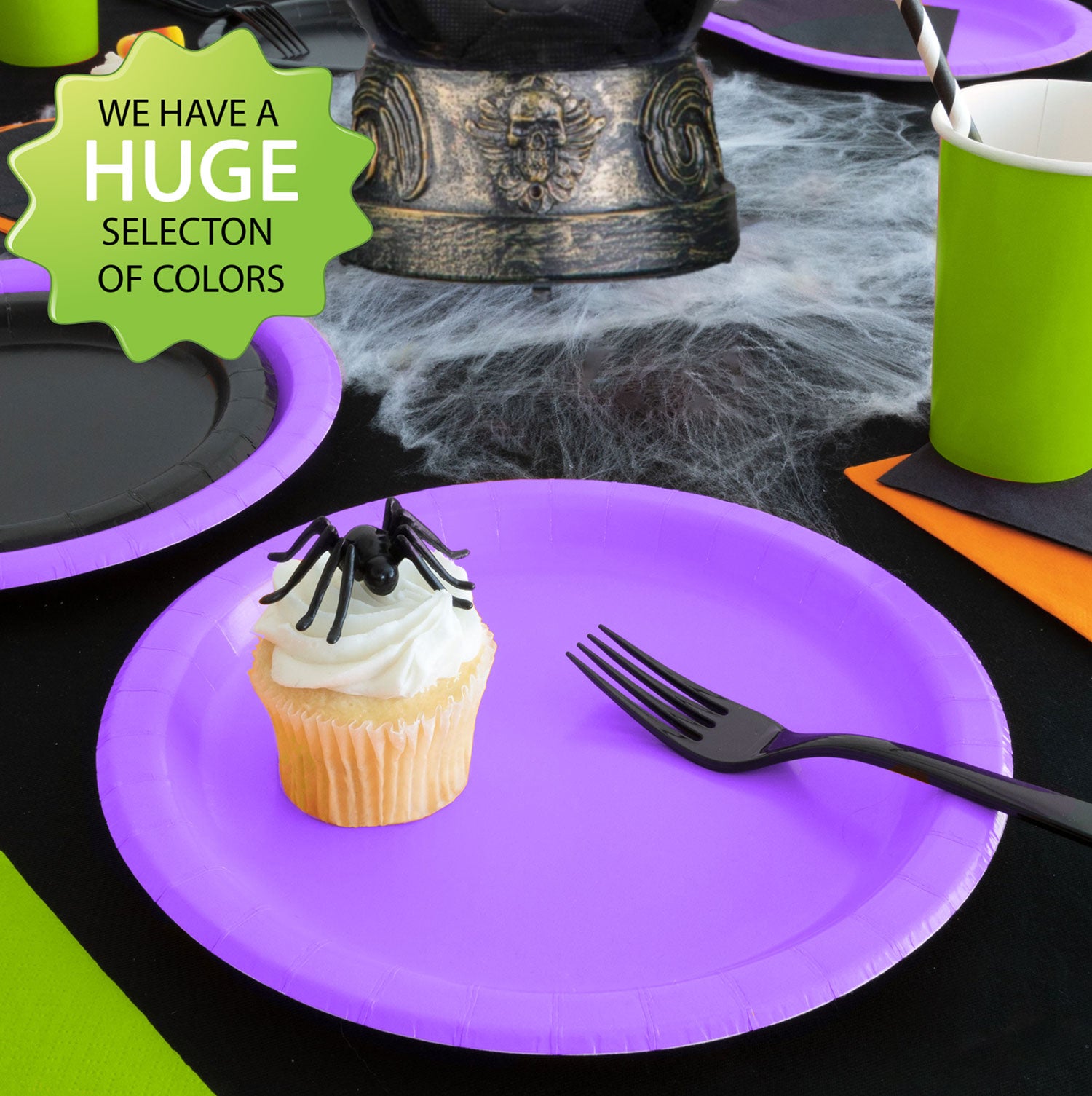 A picture of a Halloween table setting featuring solid color tableware in Halloween colors. in the foreground is a purple paper plate with a black fork and a cupcake with a spider decoration on it.