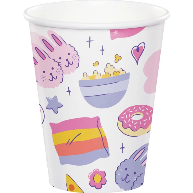 A white paper party cup decorated with a colorful, playful pattern of pink bunnies, a bowl of popcorn, a pink donut, a rainbow-striped pillow, and small stars and hearts.