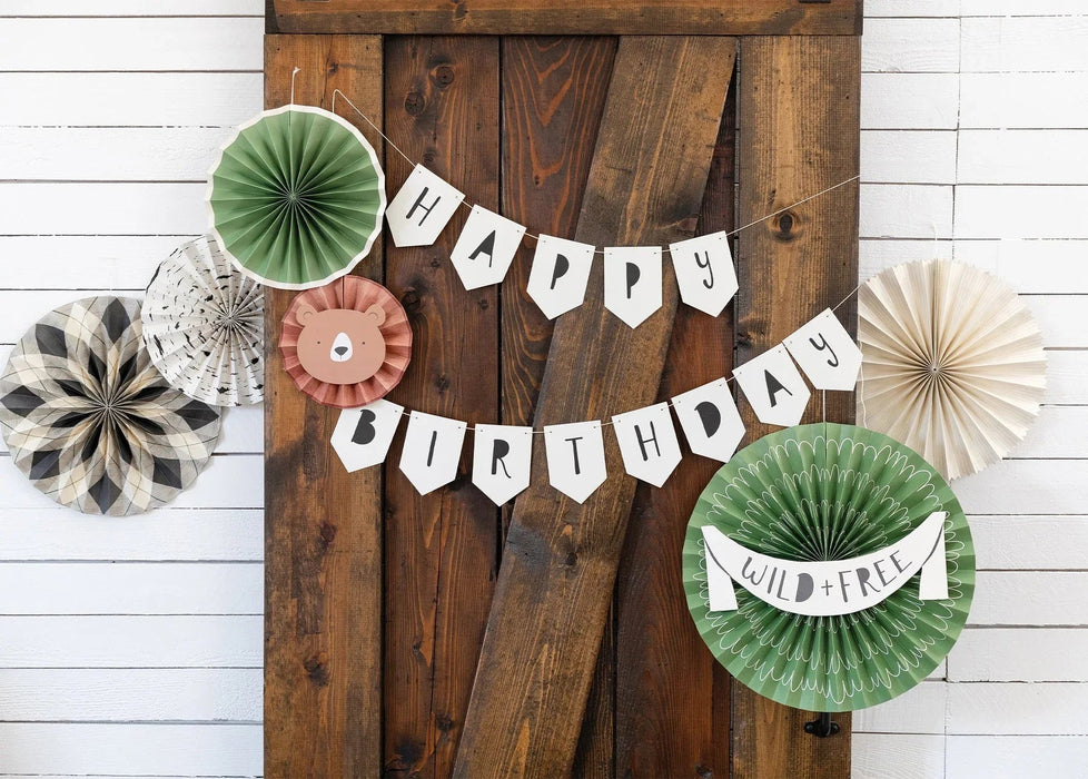 Happy Birthday banner with white pennant flags and a "Wild+Free" label, accompanied by decorative paper fan rosettes in green, cream, and patterned designs arranged on rustic wood and white shiplap backgrounds.