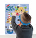 Toddler from behind wearing a blue polka dot collar and gray sweater, placing red X stickers on a pirate-themed game board featuring a treasure map, pirate ship, lighthouse, and "Ahoy Matey" text.