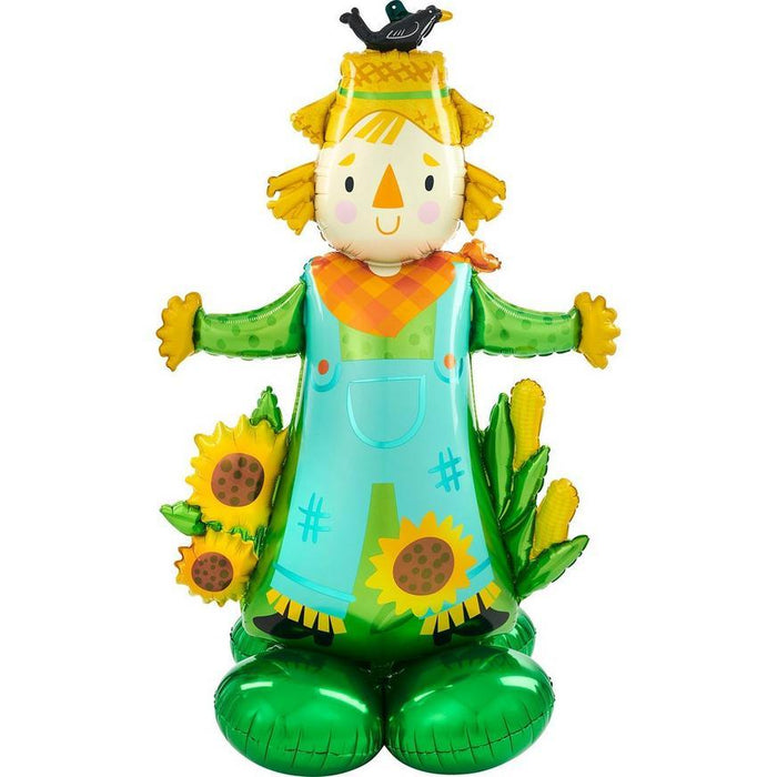 Cheerful scarecrow-shaped foil balloon with yellow straw hair, triangular orange nose, rosy cheeks, turquoise overalls decorated with sunflowers, orange neckerchief, outstretched green arms with yellow mittens, and green base stand with a small black bird perched on top.