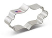 Stainless steel cookie cutter with an ornate, elongated plaque shape featuring wavy scalloped edges and a small colorful label on the upper surface.