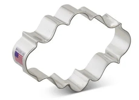 Silver metal oval plaque-shaped cookie cutter with wavy scalloped edges and a small American flag label on the side.