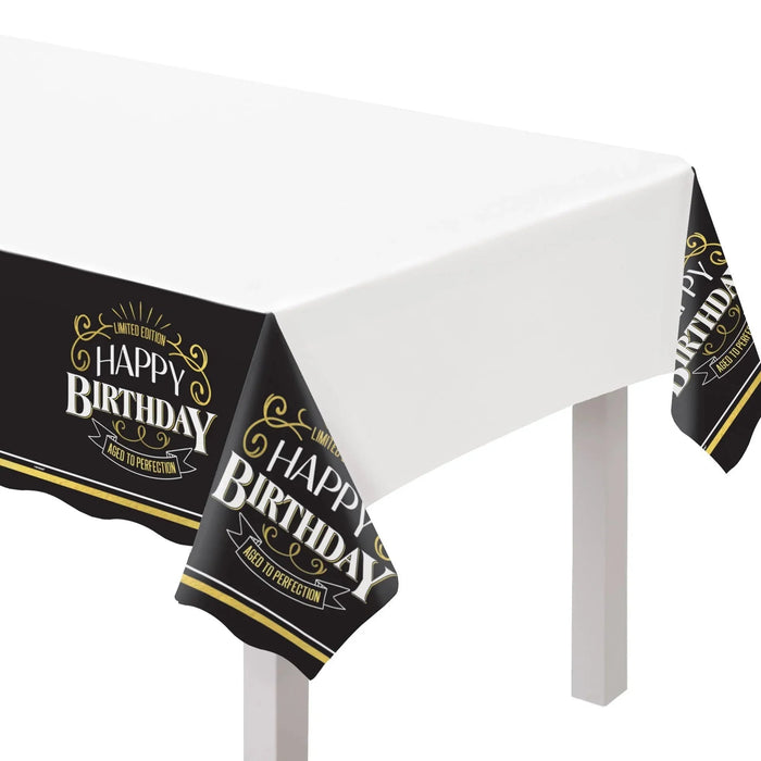 Better With Age Birthday plastic table cover measuring 54 x 102 inches, featuring a white center with black side panels decorated with gold accents and repeating Happy Birthday designs.