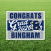 Navy and gray yard sign with white text reading "Congrats Grad 2026 Bingham" and a decorative graduation cap graphic, mounted on a green grass background.