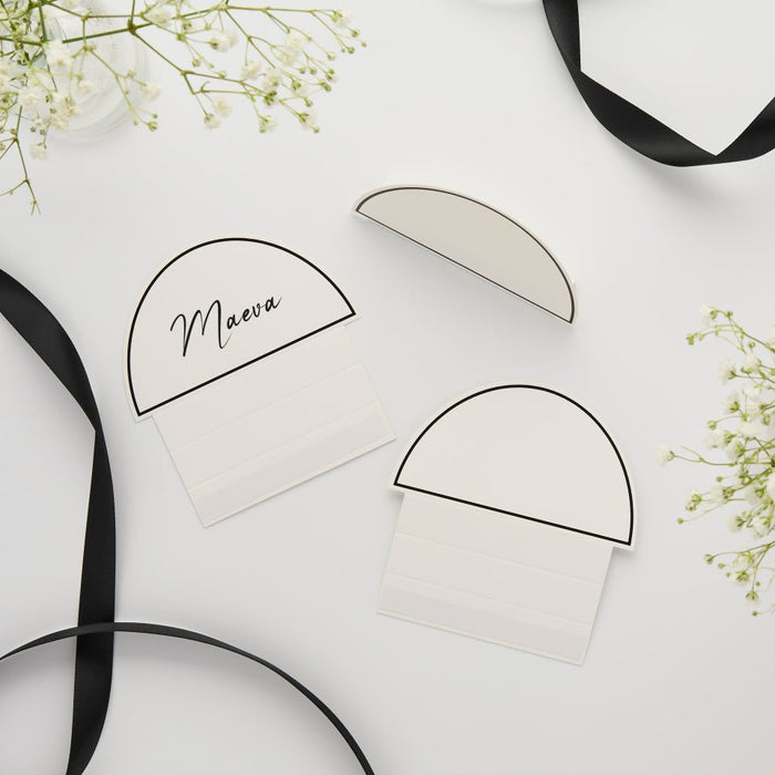 Three cream-colored tented place cards with black borders arranged on a white surface, one labeled "Maeva," surrounded by black ribbons and baby's breath flowers.