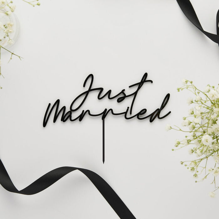 Black acrylic cake topper with "Just Married" script text and a pointed base, displayed on white background with delicate baby's breath flowers and black ribbon accents.