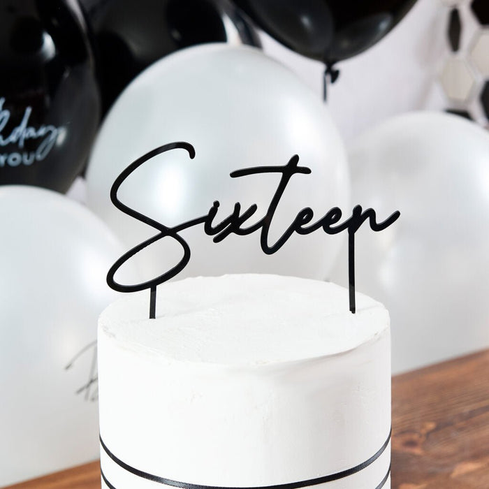 Black wire cake topper reading "Sixteen" inserted into a white frosted cake decorated with a black ribbon band and white balloons in the background.