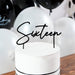 Black wire cake topper reading "Sixteen" inserted into a white frosted cake decorated with a black ribbon band and white balloons in the background.