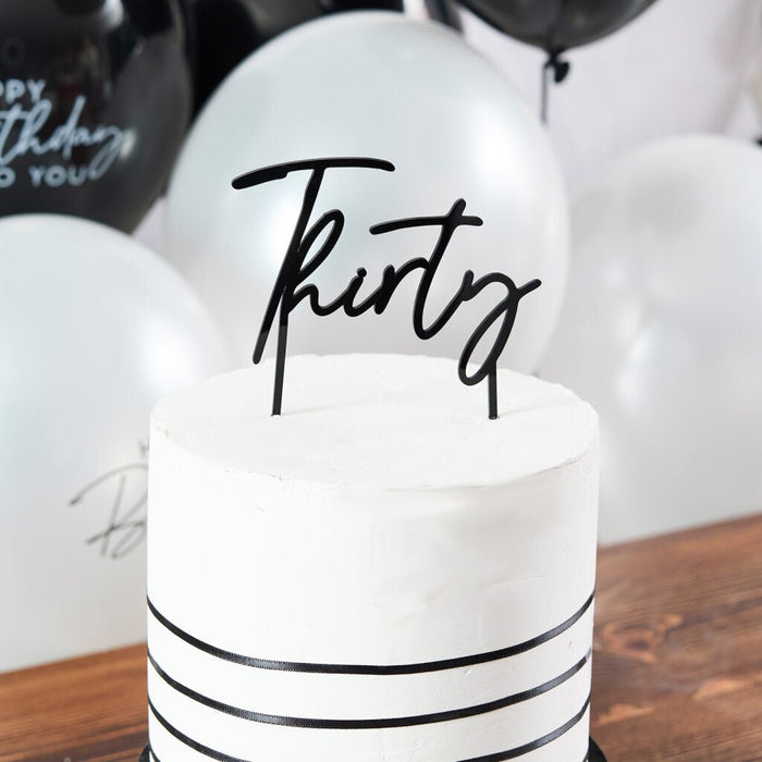 White round cake with black ribbon stripes and a black cursive "thirty" topper, surrounded by white and black balloons on a wooden surface.