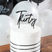 White round cake with black ribbon stripes and a black cursive "thirty" topper, surrounded by white and black balloons on a wooden surface.