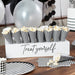 White display stand with "Treat yourself" text holds eight black and white striped popcorn cones filled with popcorn, arranged in a row on a wooden table.
