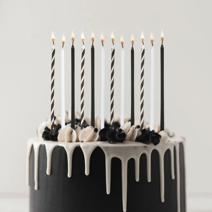 Black and white striped and solid tall birthday candles with lit flames arranged in a black cake with white drip frosting and black and white frosting piped decorations.