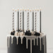 Black and white striped and solid tall birthday candles with lit flames arranged in a black cake with white drip frosting and black and white frosting piped decorations.