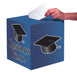 Blue cubic card box with "Congrats Grad" text and graduation cap graphics, shown with a hand inserting a white card into the top slot.
