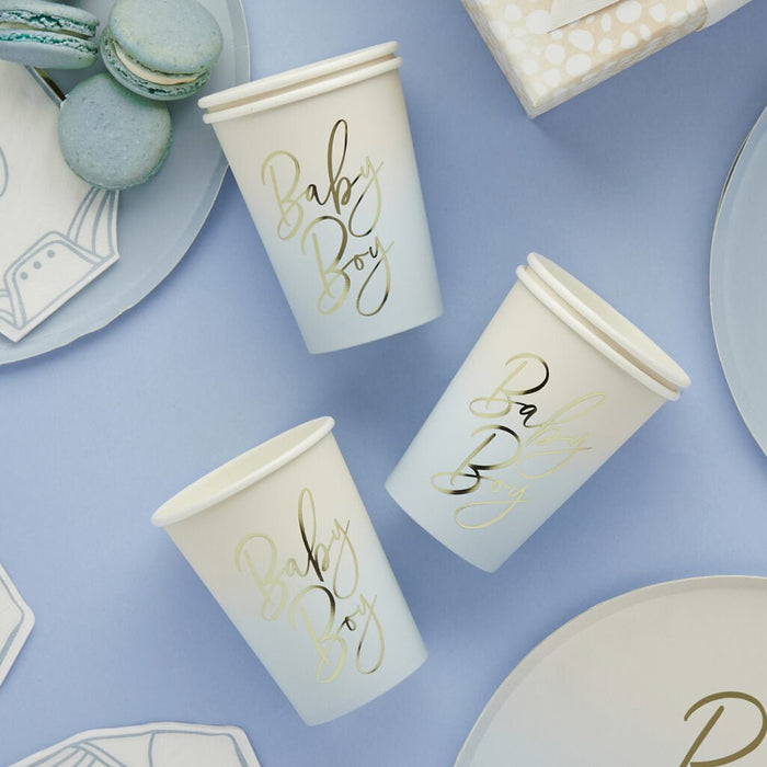 Three cream-colored paper cups with gold script text reading "Baby Boy" arranged on a blue background alongside matching plates, macarons, and napkins.