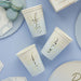 Three cream-colored paper cups with gold script text reading "Baby Boy" arranged on a blue background alongside matching plates, macarons, and napkins.