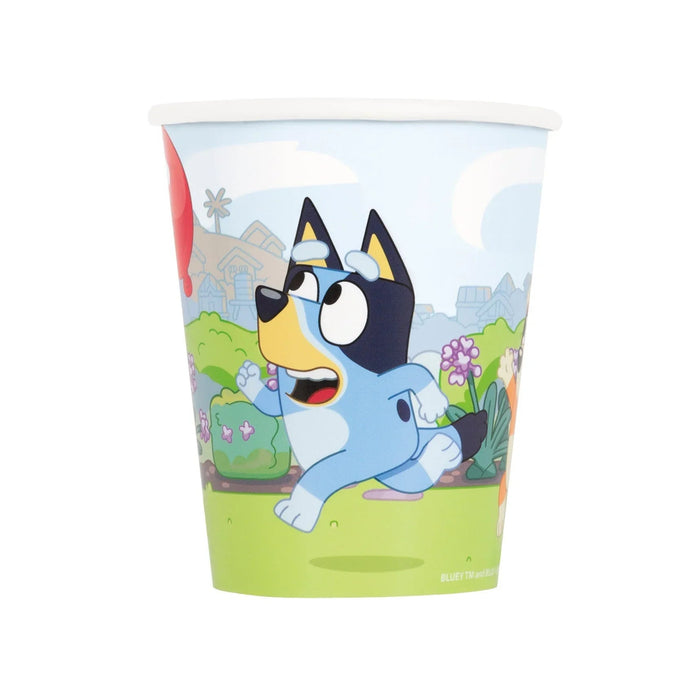 Disposable paper cup featuring Bluey the animated dog character in light blue with black ears and yellow accents, set in a cheerful garden scene with green grass, purple flowers, and blue sky.