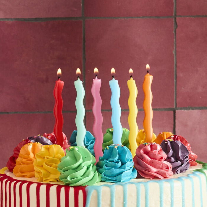 Colorful birthday cake decorated with six wavy spiral candles in red, white, pink, light blue, yellow, and orange, topped with piped frosting flowers in matching rainbow colors on a striped fondant base.