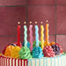 Colorful birthday cake decorated with six wavy spiral candles in red, white, pink, light blue, yellow, and orange, topped with piped frosting flowers in matching rainbow colors on a striped fondant base.