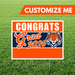Orange and navy yard sign with "CONGRATS Grad 2026" text and a tiger mascot head, displayed on grass with a white border.