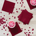 Burgundy paper napkins arranged on a white wooden surface with matching burgundy confetti circles and two white bowls containing pink dessert or punch.