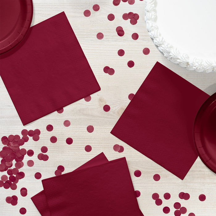Burgundy paper napkins and coordinating party supplies arranged on a white wooden surface, including stacked napkins, decorative confetti circles, and a white ruffled plate.