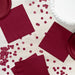 Burgundy paper napkins and coordinating party supplies arranged on a white wooden surface, including stacked napkins, decorative confetti circles, and a white ruffled plate.